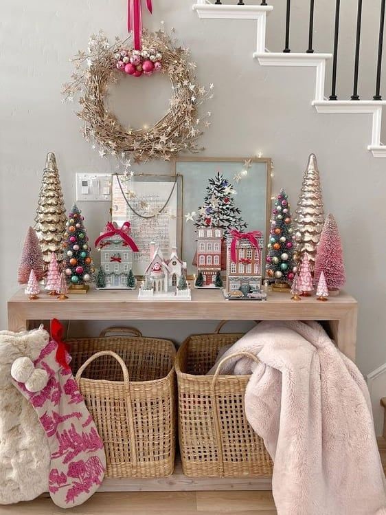 Pink Christmas entryway decorated with bottle-brush trees, festive village houses, metallic trees, and a blush wreath above a console table.