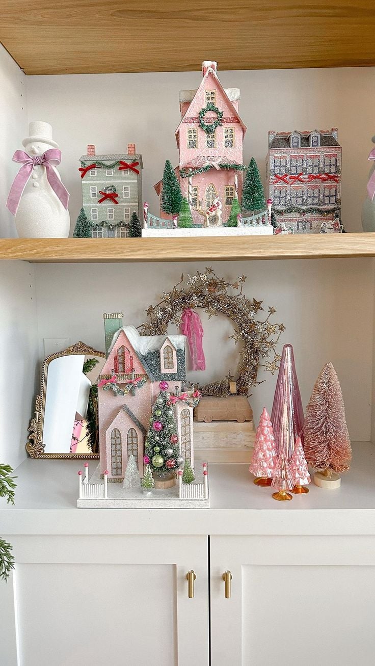 Shelf styled with pink Christmas village houses, bottle-brush trees, a gold wreath, and festive pastel holiday decor.