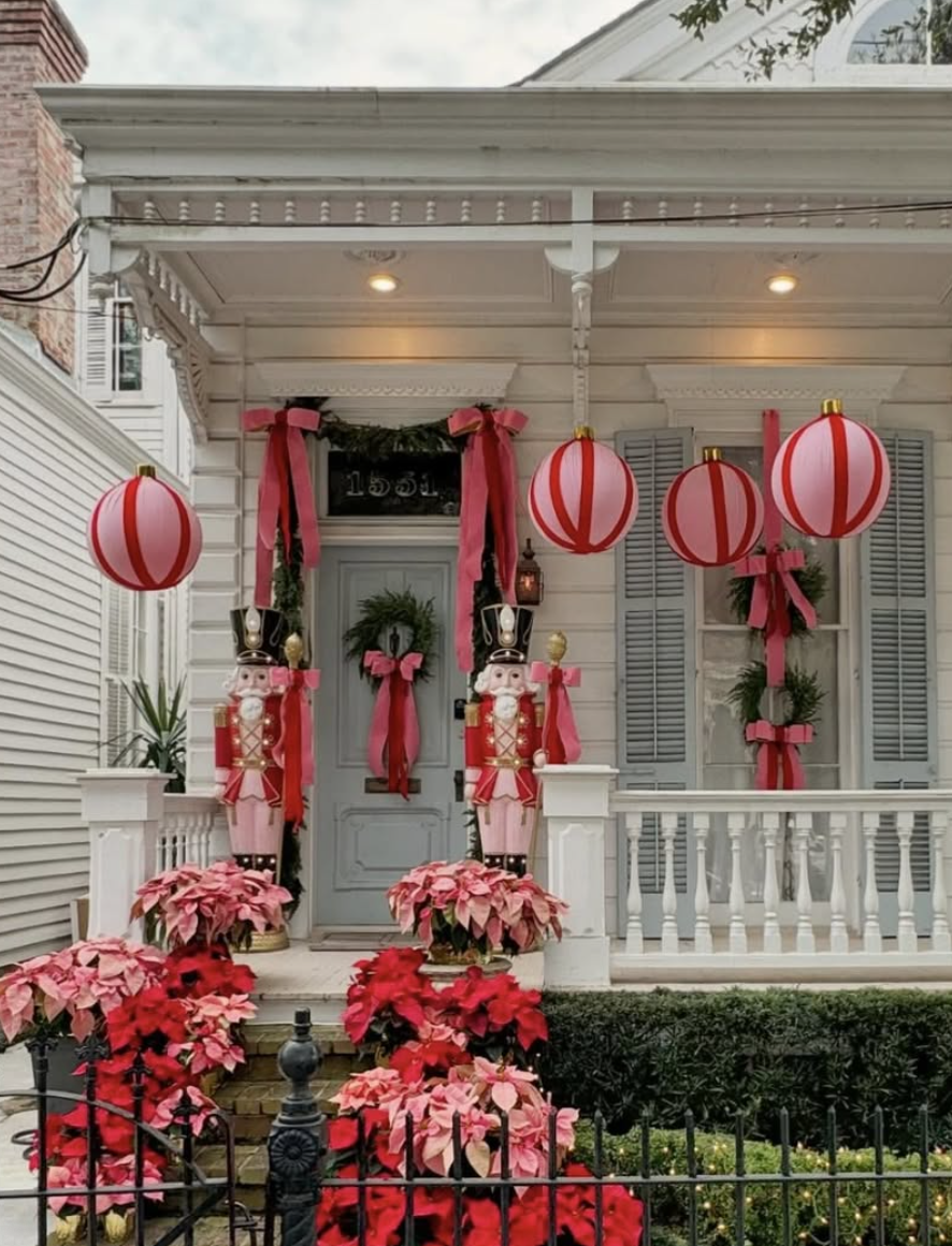 Festive Christmas front porch decorated with oversized pink nutcrackers, red-and-white hanging ornaments, wreaths, pink bows, and poinsettias.