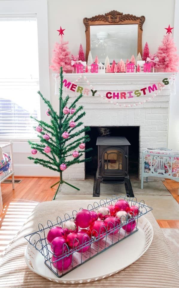 Bright pink Christmas mantel with mini village houses, bottle-brush trees, a colorful Merry Christmas banner, and a small retro-style tree decorated with pink ornaments.