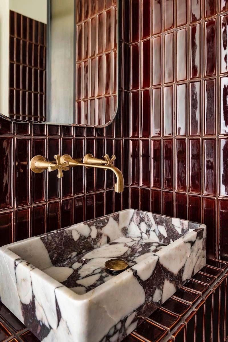 A modern bathroom featuring glossy deep red vertical tile, a brass wall-mounted faucet, and a square red-and-white marble sink.