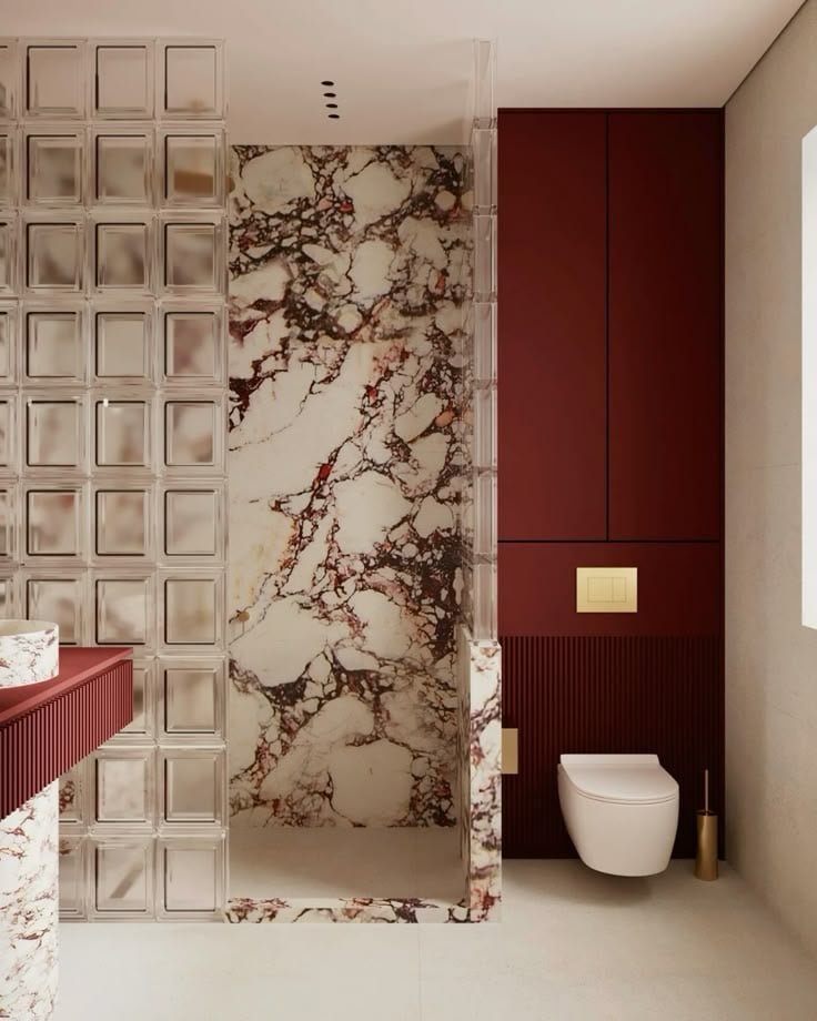 A contemporary bathroom featuring a red and white marble shower, a glass block partition wall, burgundy cabinetry, and brass accents.