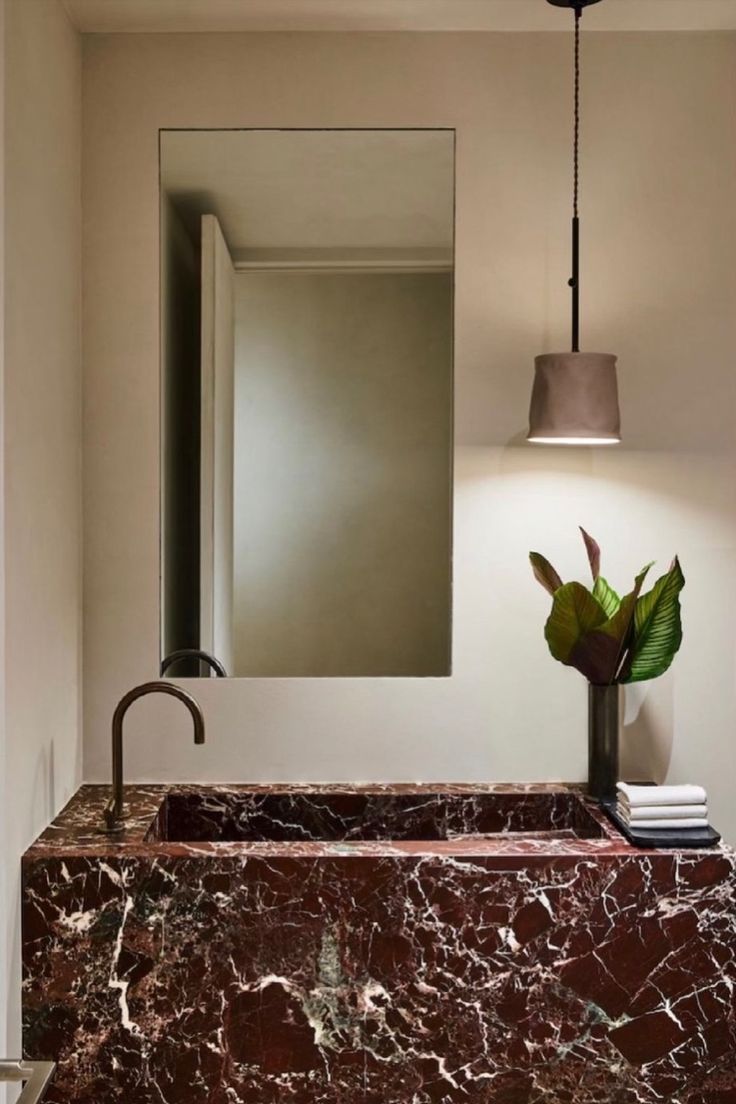 A minimalist bathroom featuring a large red marble vanity with dramatic white veining, a curved bronze faucet, a simple rectangular mirror, and a modern pendant light.