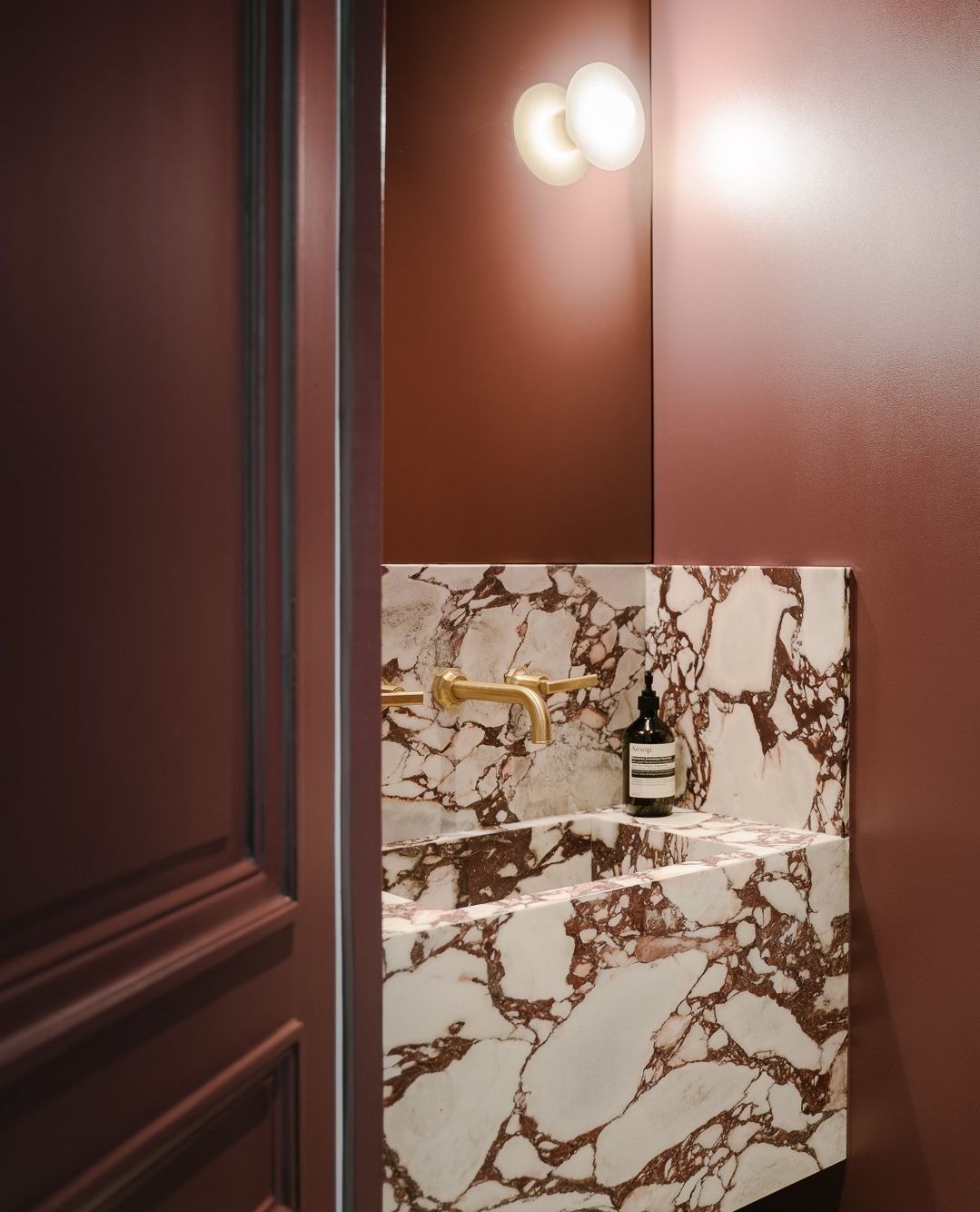 A stylish powder room featuring a bold red-and-white marble vanity with a brass wall-mounted faucet, warm red painted walls, and a modern round wall sconce.