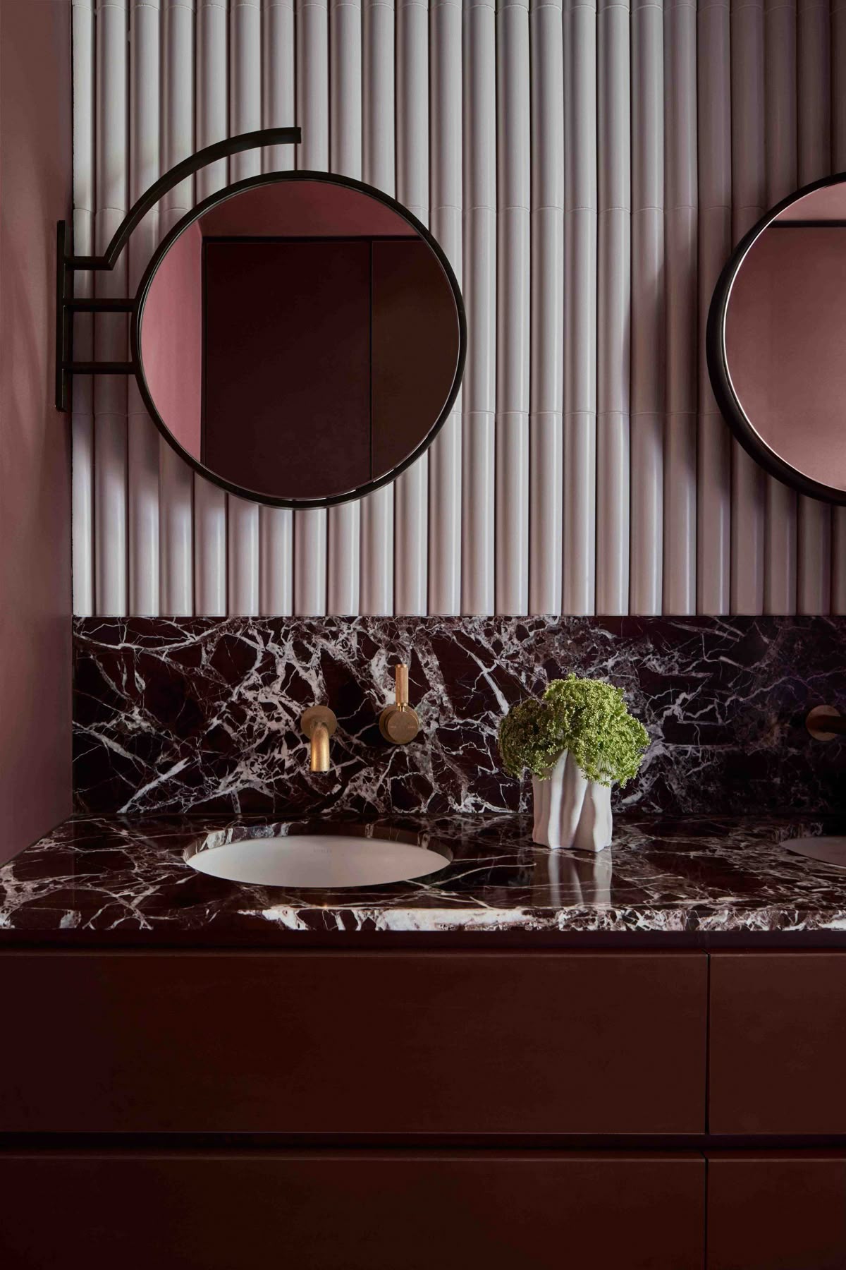 A stylish bathroom featuring a deep red marble vanity with bold white veining, round black-framed mirrors, brass wall-mounted faucets, and fluted white wall tile.