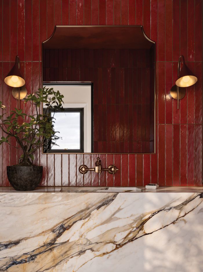 A bold bathroom featuring glossy red vertical tile, a sculptural white marble vanity with gold and charcoal veining, brass wall-mounted faucets, and modern red sconces.