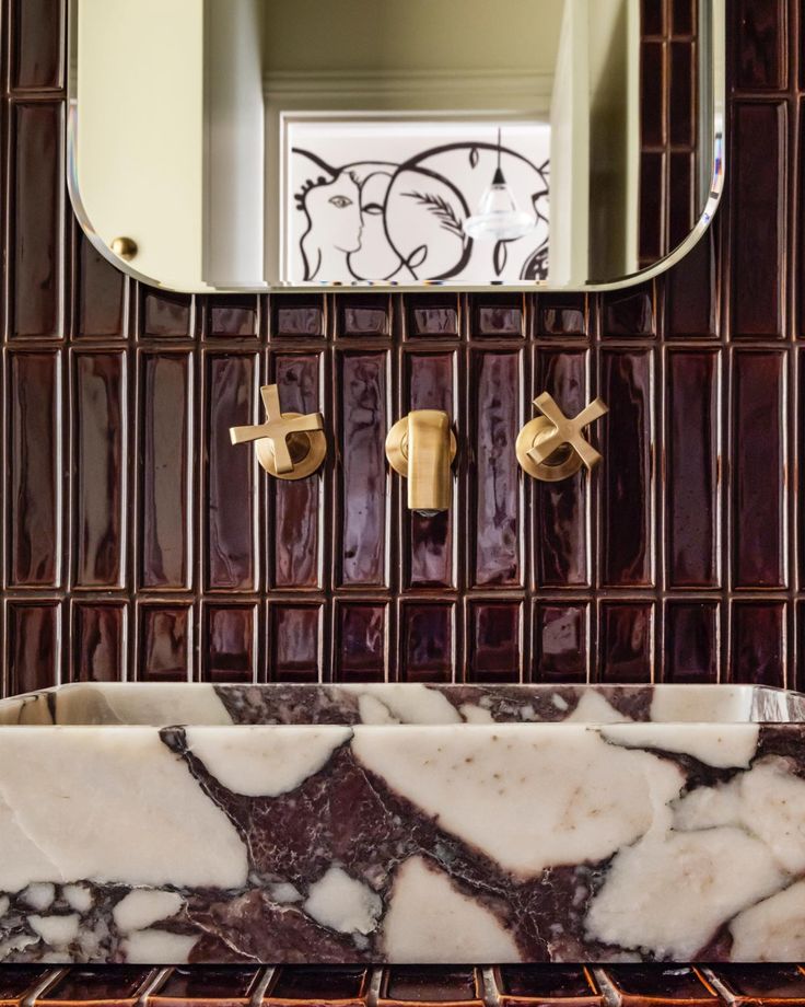 A modern bathroom featuring glossy deep red tile, a sculptural red-and-white marble vessel sink, and brass wall-mounted faucets beneath a rounded-edge mirror.
