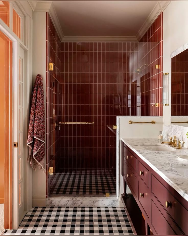 A classic bathroom featuring glossy red vertical tile in the shower, a red wood vanity with a marble countertop, brass fixtures, and a black-and-white checkerboard floor.