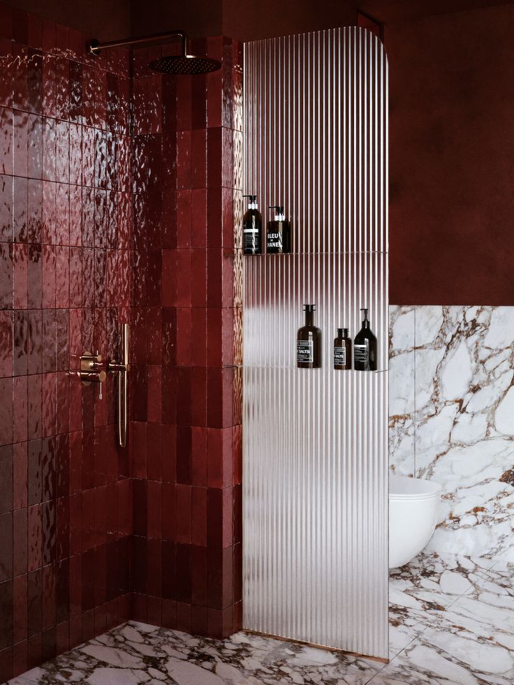 A modern bathroom featuring a deep red tile shower, fluted glass divider with built-in shelves, brass shower fixtures, and red-and-white marble flooring.