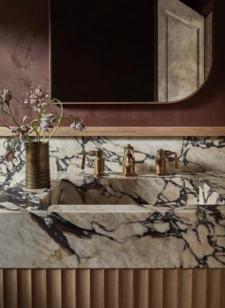 A luxurious bathroom vanity featuring dramatic red-veined marble, brass faucet hardware, a fluted wood cabinet base, a rounded rectangular mirror, and a rustic red plaster wall.