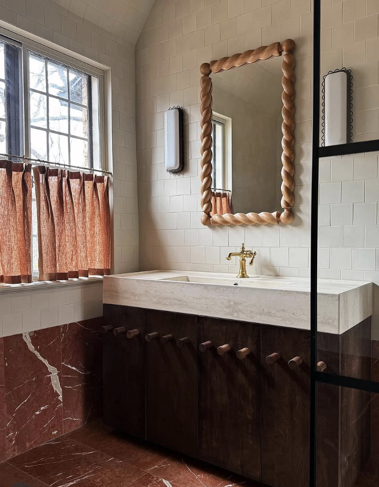 A charming bathroom featuring red marble flooring and wainscoting, dark wood cabinetry, a cream stone trough sink, brass faucet, twisted-frame mirror, and arched white tile walls.