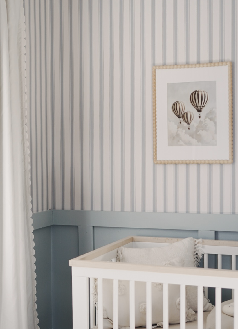 Baby nursery with blue striped wallpaper, dusty blue board and batten, white crib, and framed hot air balloon artwork