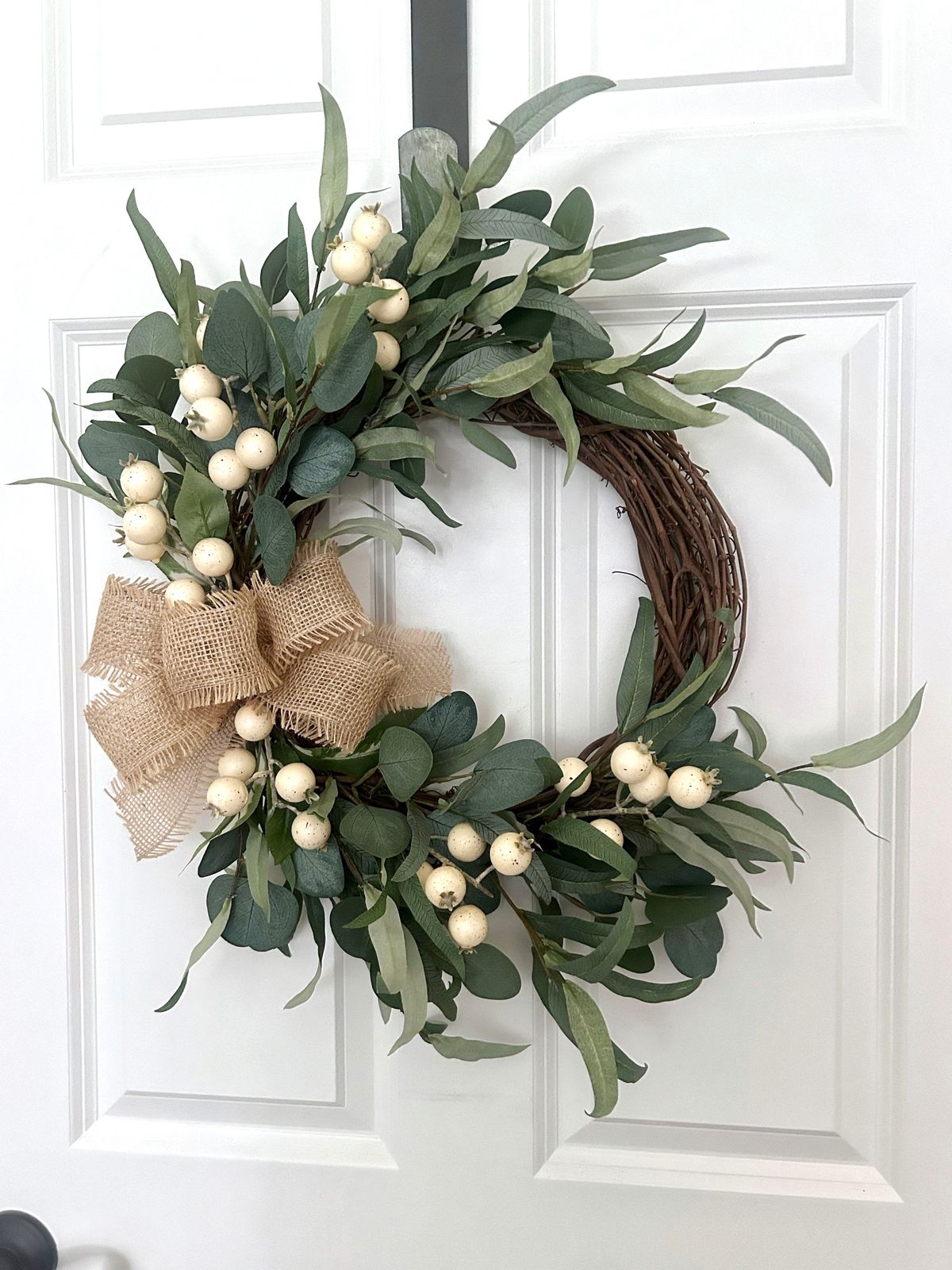 Farmhouse eucalyptus wreath with white berries and a rustic burlap bow hanging on a white paneled front door