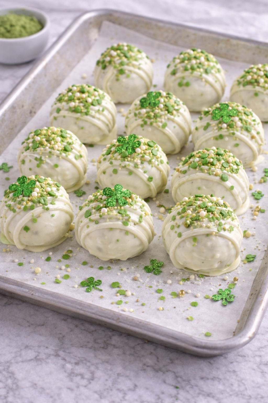 Healthy St. Patrick’s Day matcha truffles coated in white chocolate and decorated with green sprinkles and shamrocks, setting on a parchment-lined baking sheet.