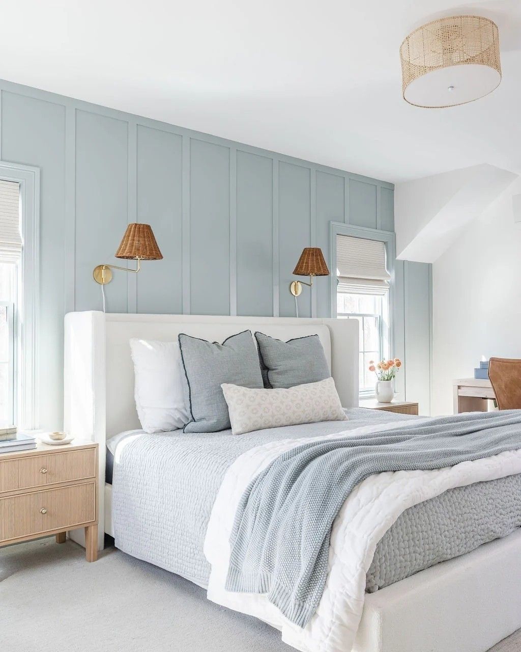 Light blue bedroom with board and batten accent wall, white upholstered bed, layered blue bedding, and woven light fixtures