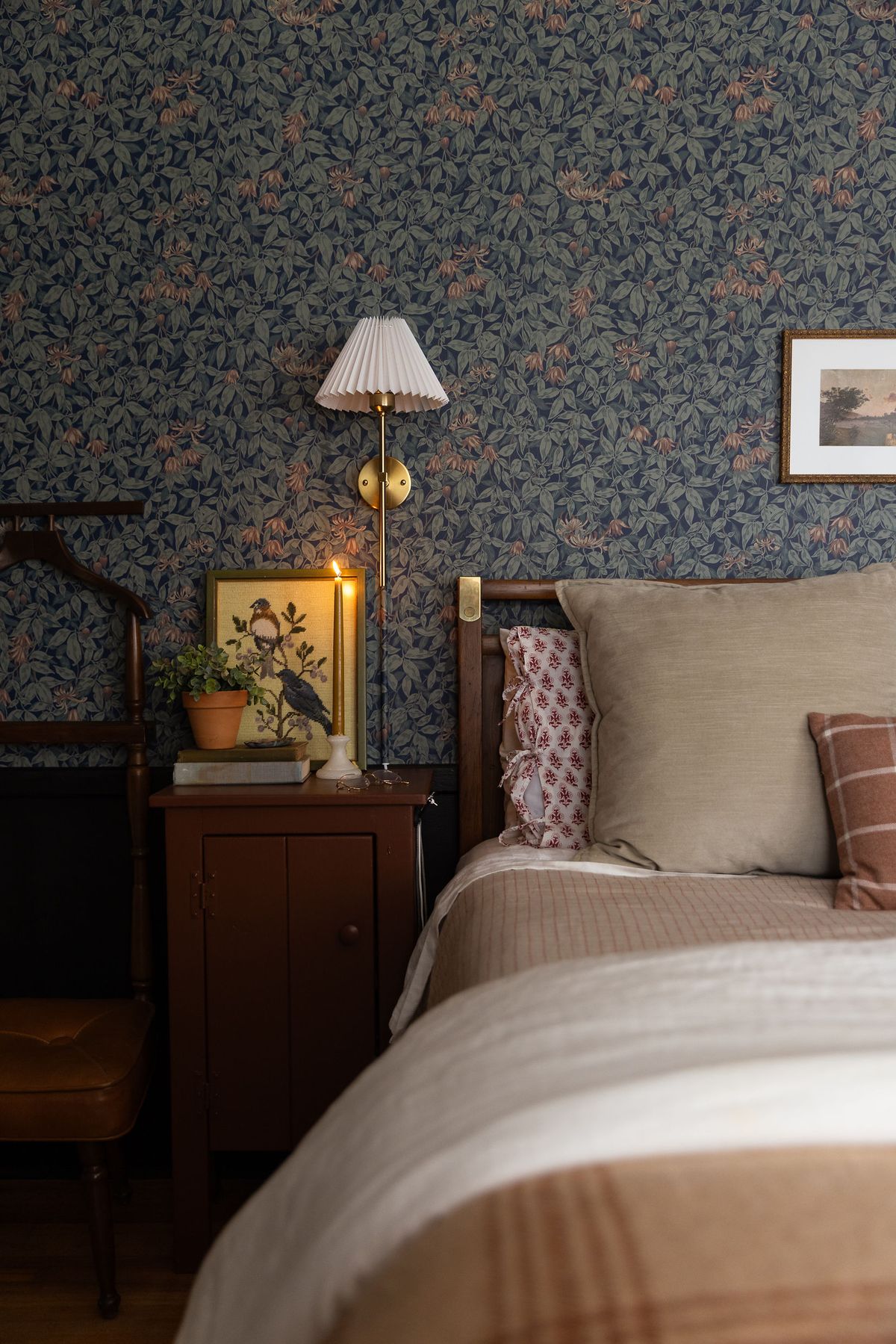 Cozy bedroom with dark blue botanical wallpaper, wooden bed frame, vintage nightstand, and warm brass wall sconce