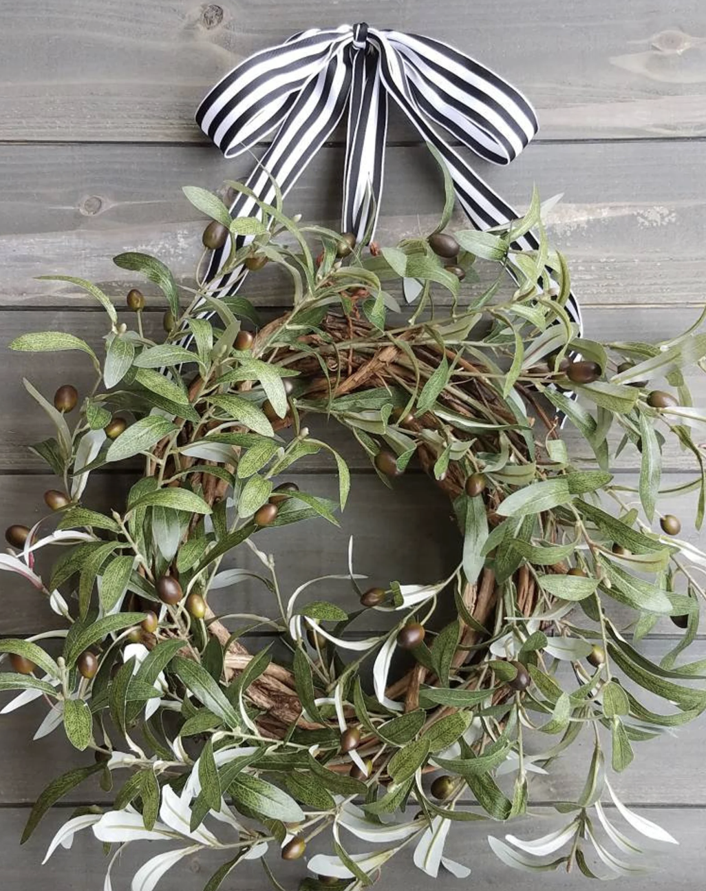 Olive branch wreath with green leaves and olives, finished with a black and white striped bow on a rustic wooden background