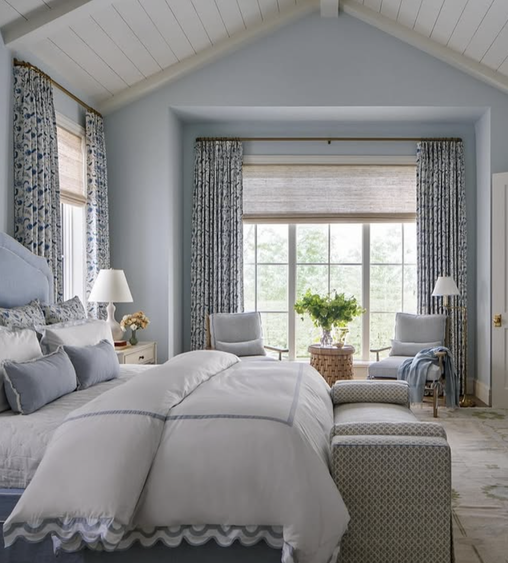 Soft blue bedroom with large windows, patterned blue curtains, white bedding, upholstered bench, and cozy seating area