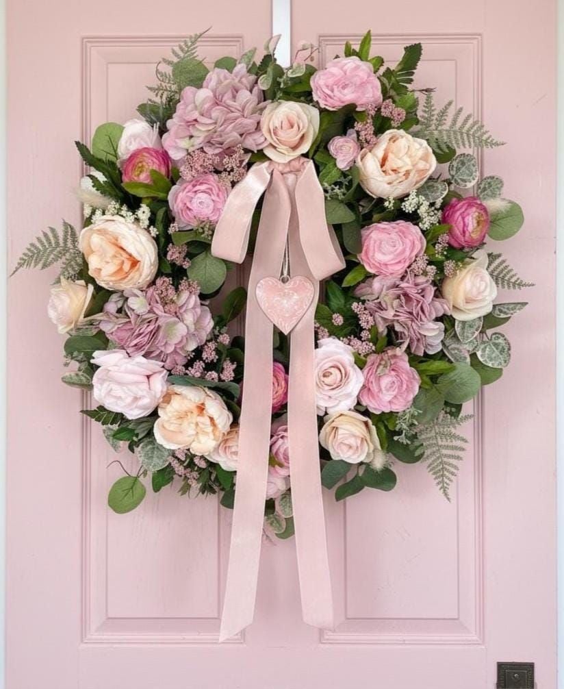Whimsical pink and blush floral wreath with roses, greenery, and a long ribbon with a hanging heart on a soft pink front door