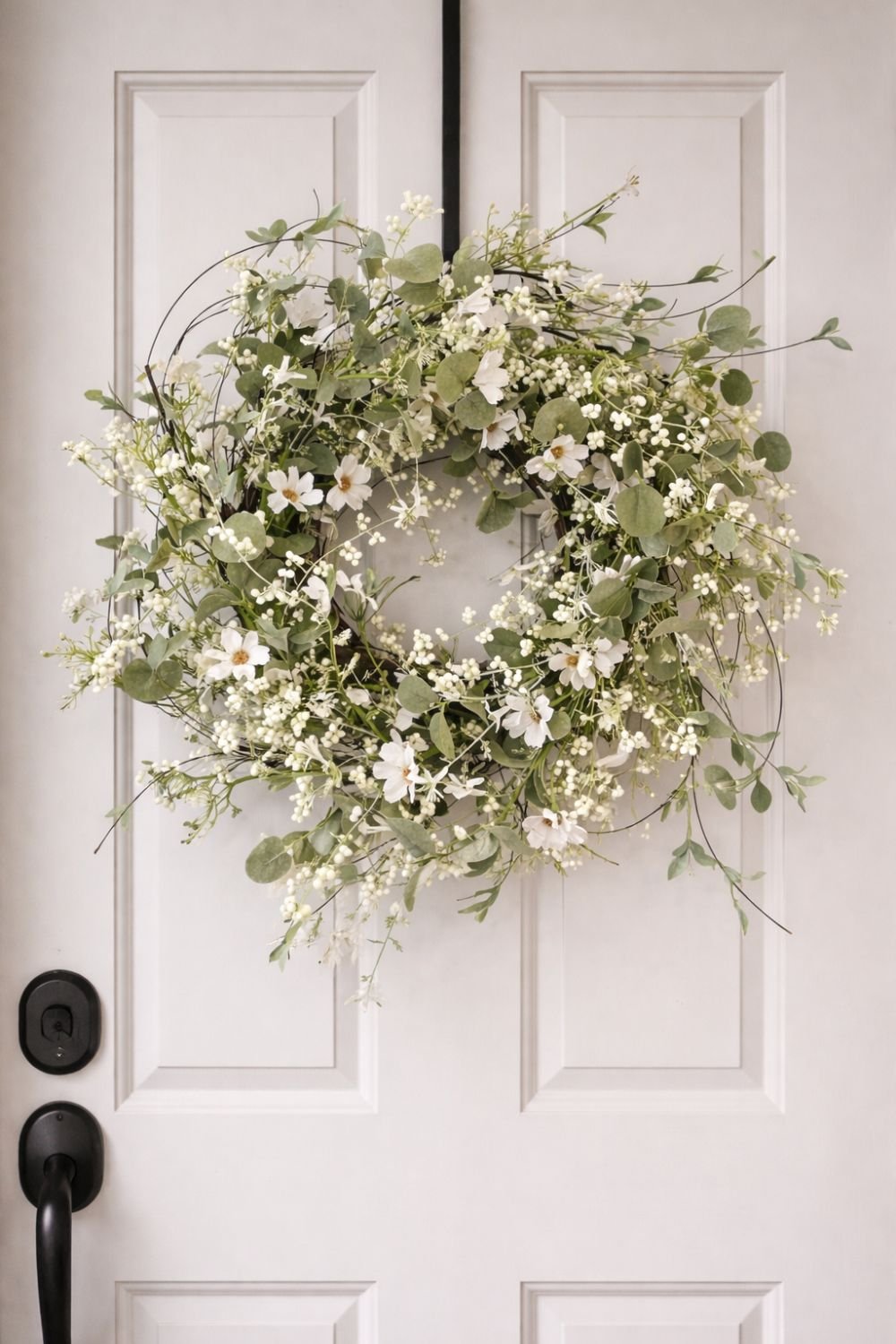 Delicate white floral and eucalyptus spring wreath hanging on a light-colored door with a black door handle