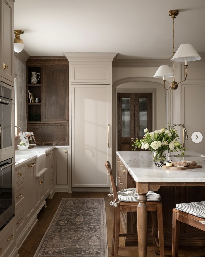 Farmhouse kitchen with a marble island, wooden cabinetry, brass lighting, and upholstered island chairs.