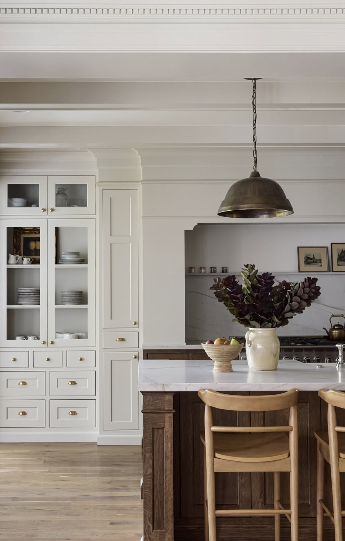 White farmhouse kitchen with glass-front cabinets, a marble island, wooden bar stools, and a brass pendant light.