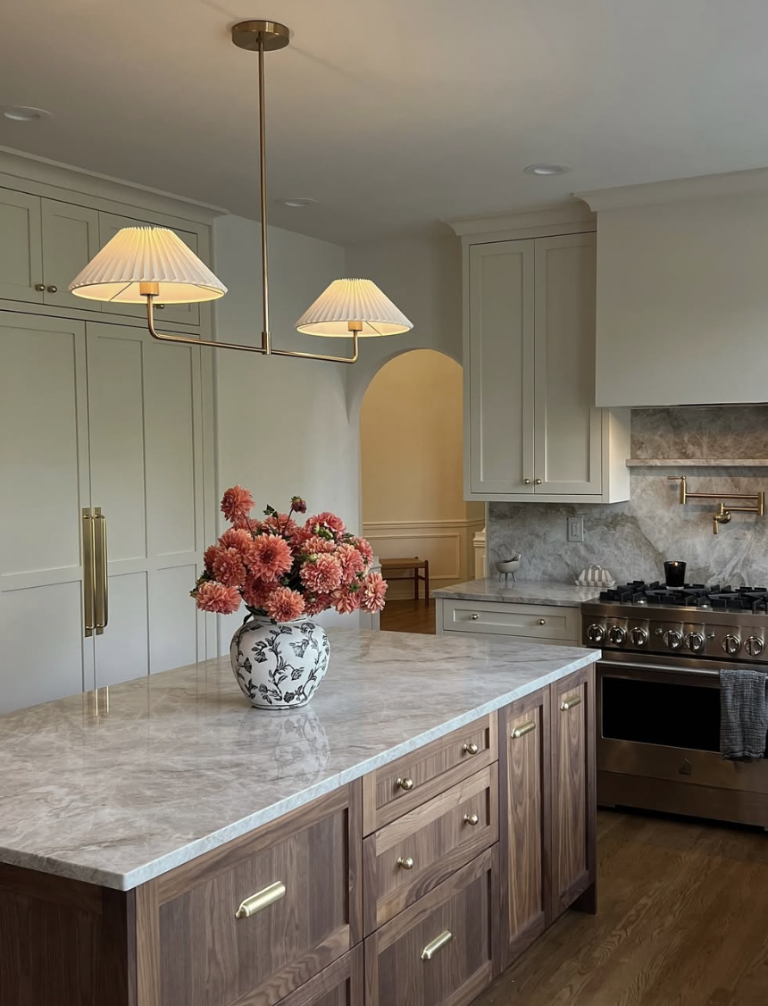 Kitchen with a marble island, walnut cabinetry, brass hardware, and soft pendant lighting above a floral centerpiece.