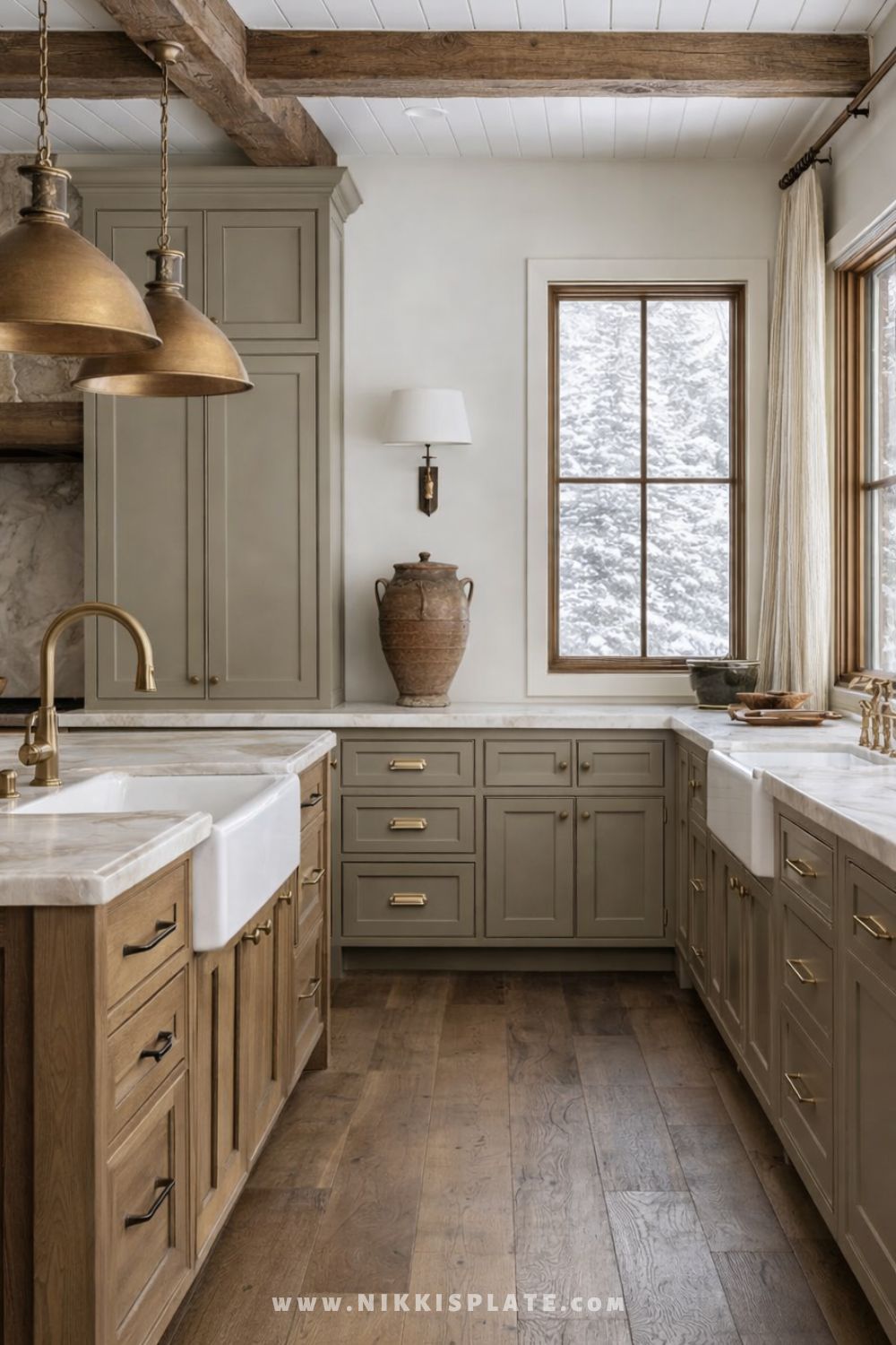 Aged brass farmhouse pendant lights hanging above a kitchen island with a white apron-front sink and rustic wood beams.