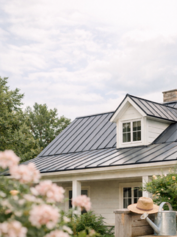 Modern farmhouse with a black metal roof surrounded by soft greenery and flowers