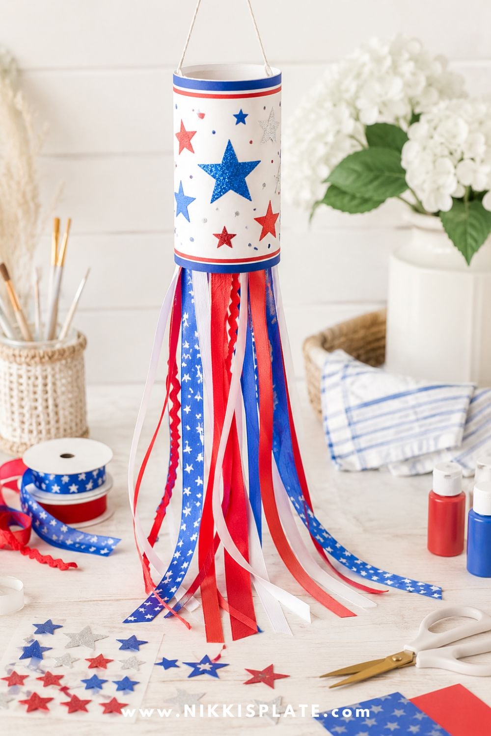 4th of July windsock craft for kids with red, white, and blue ribbons, star stickers, paint, and craft supplies on a light table