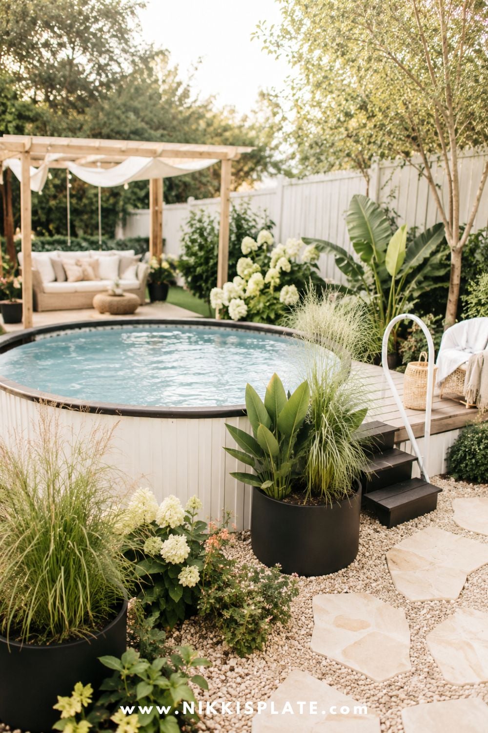 Above ground pool with modern black planters, tropical greenery, ornamental grasses, white hydrangeas, and a bright gravel walkway.