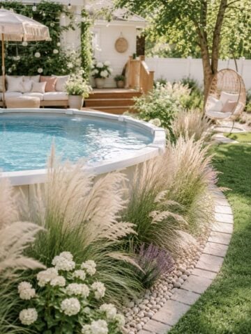 Above ground pool with soft ornamental grasses, white flowers, river rock edging, and a cozy backyard lounge area.