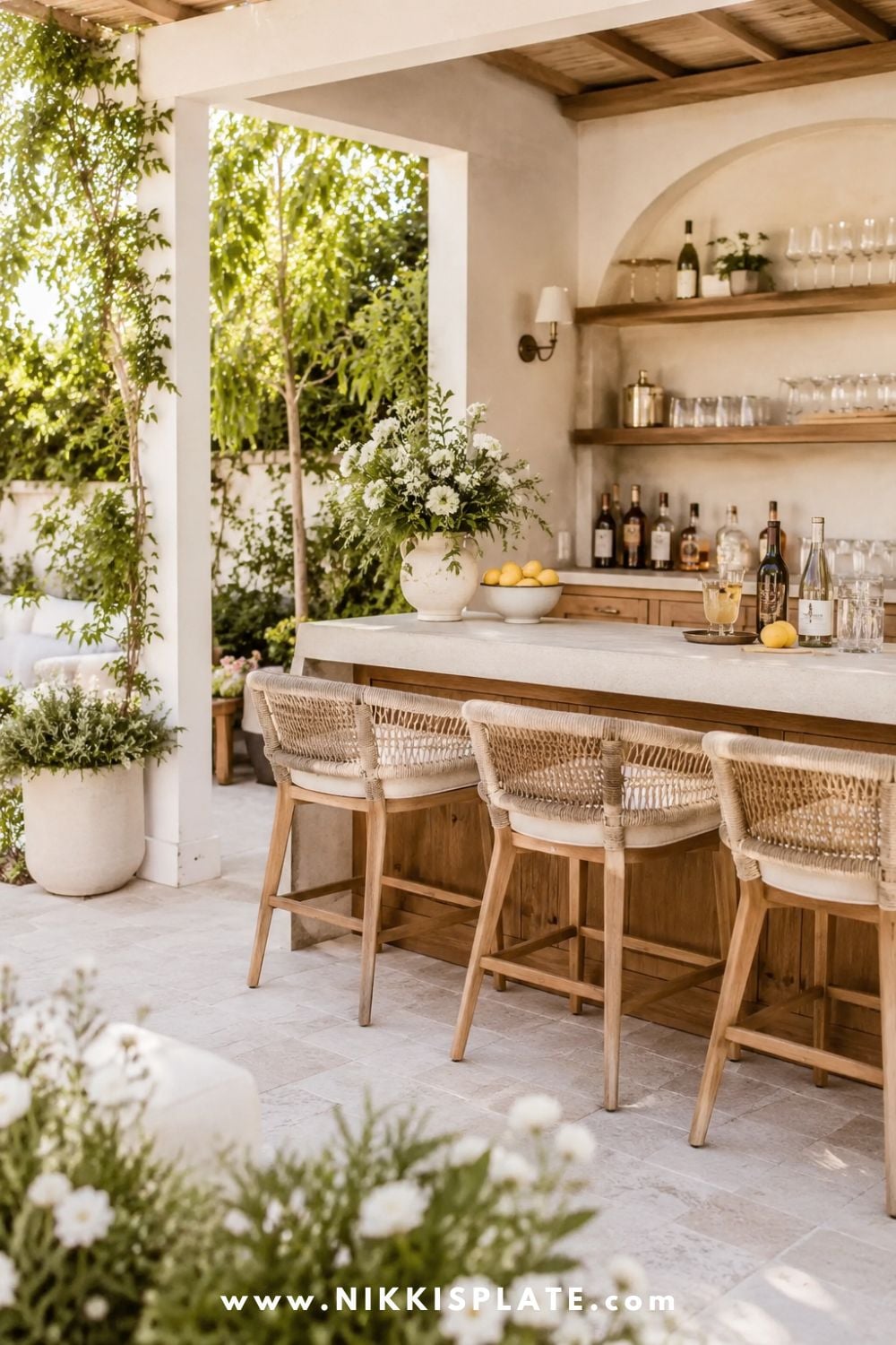 Built-in backyard bar with warm wood cabinetry, light stone countertop, wicker bar stools, open shelves, soft flowers, greenery, and airy farmhouse patio decor.