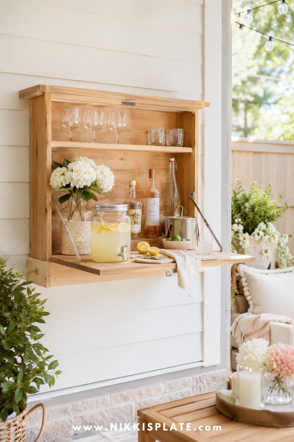 Fold-down wall bar with warm wood shelves, lemonade dispenser, glassware, soft flowers, citrus, and cozy farmhouse patio decor