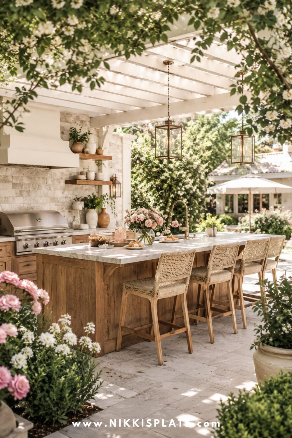Outdoor kitchen with bar seating, warm wood island, wicker bar stools, white pergola, stone backsplash, soft flowers, and cozy garden patio decor