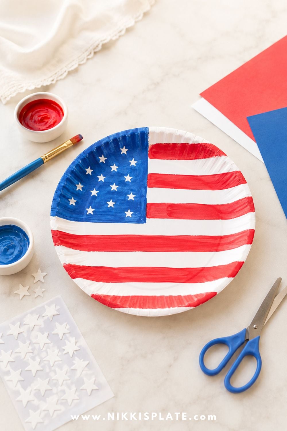 Paper plate flag craft for kids with red and white painted stripes, blue paint, and white star stickers on a craft table