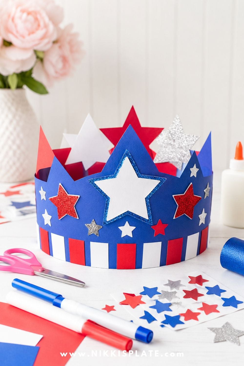 Patriotic paper crown craft for kids with red, white, and blue cardstock, star stickers, markers, scissors, and glue on a craft table