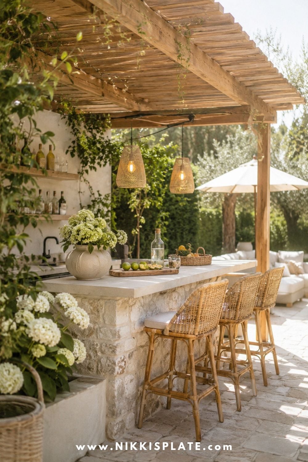 Pergola-covered bar with stone counter, warm wood beams, wicker bar stools, woven pendant lights, greenery, and cozy farmhouse patio decor