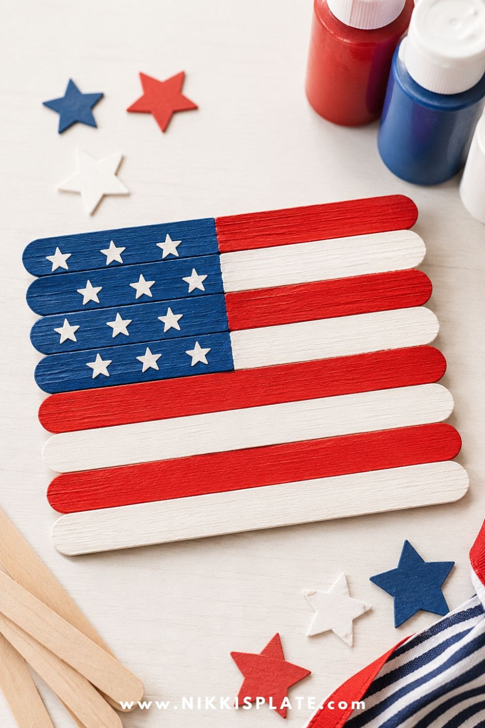 Popsicle stick American flag craft for kids with red, white, and blue painted craft sticks, star stickers, and craft supplies on a table