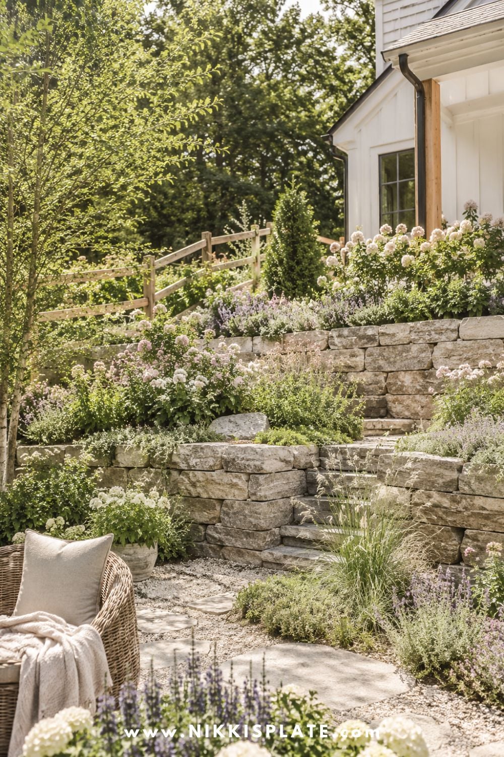 Sloped backyard landscaping with stone retaining walls, terraced garden beds, soft cottage flowers, and a cozy farmhouse-style patio path.