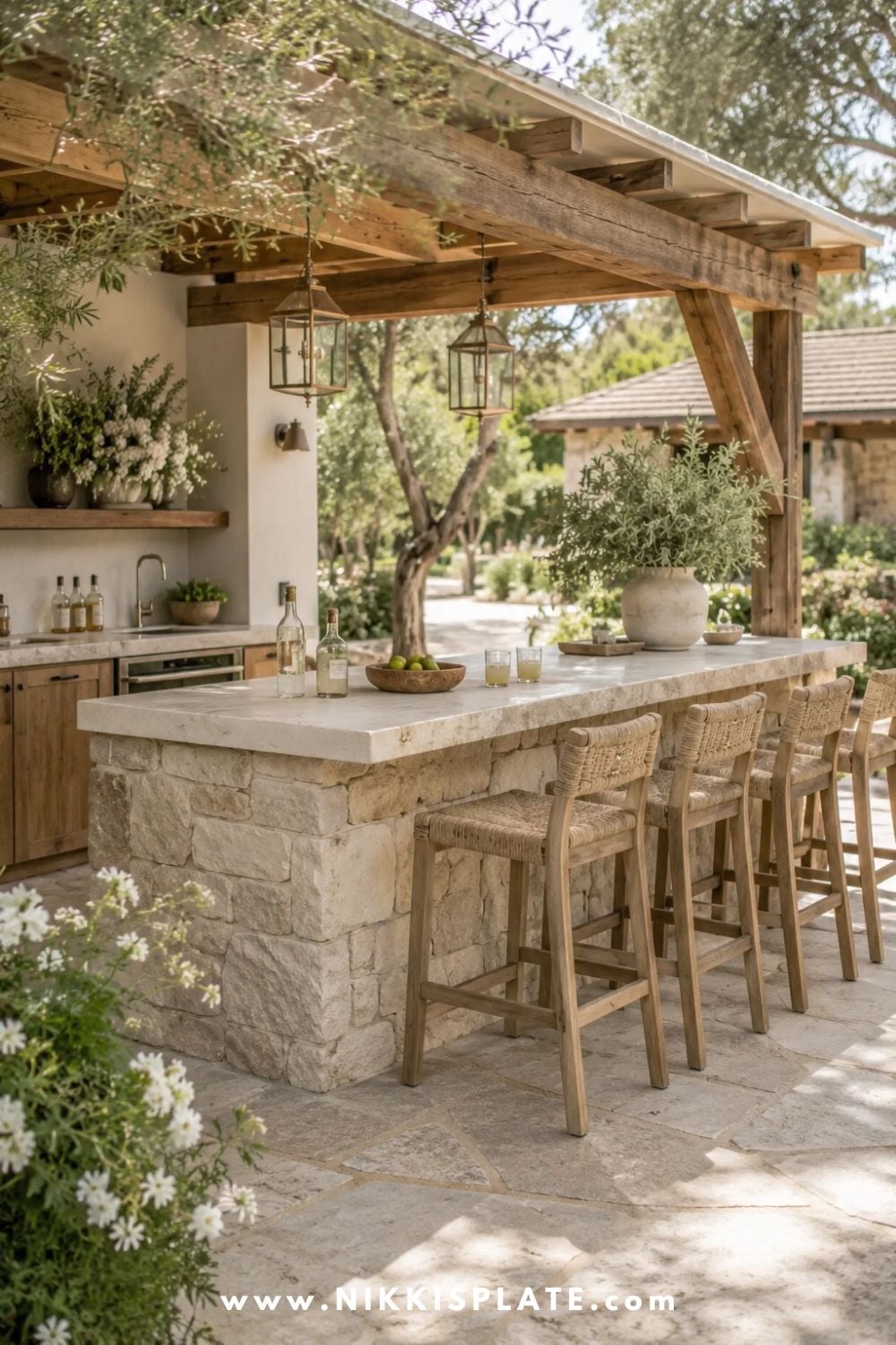 Stone outdoor bar with light stone base, warm wood pergola, woven bar stools, pale countertop, greenery, lanterns, and cozy farmhouse patio decor