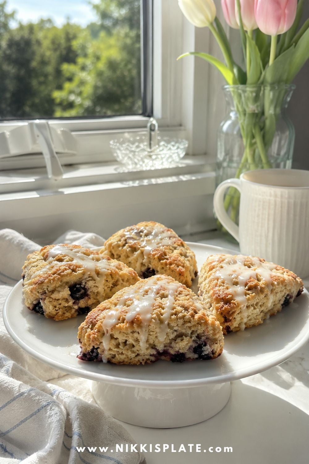 Lemon-glazed blueberry chia sourdough scones displayed on a white cake stand with tulips and a coffee mug by a sunny kitchen window.