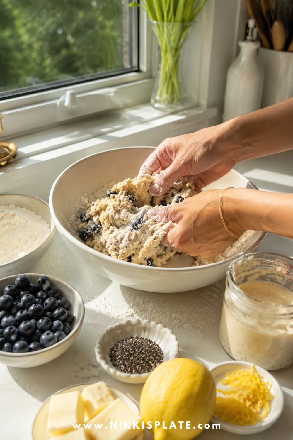 Female hands mixing blueberry chia sourdough scone dough in a white bowl with fresh blueberries, lemon, butter, chia seeds, flour, and sourdough discard nearby.