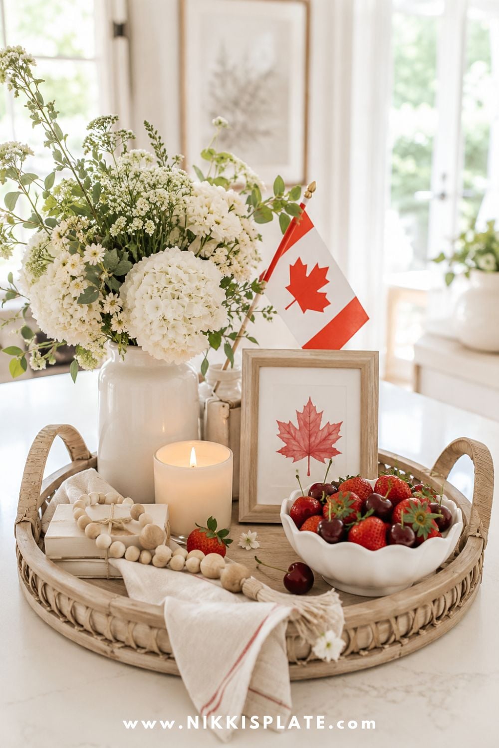 Simple Canada Day decor tray with white flowers, a mini Canadian flag, framed maple leaf printable, candle, wood beads, and a bowl of strawberries and cherries.