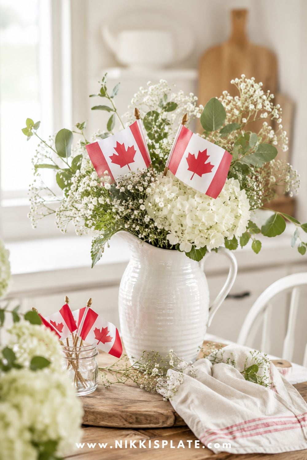 Canada Day floral centerpiece with white hydrangeas, greenery, baby's breath, and mini Canadian flags styled in a white pitcher on a rustic wooden table.