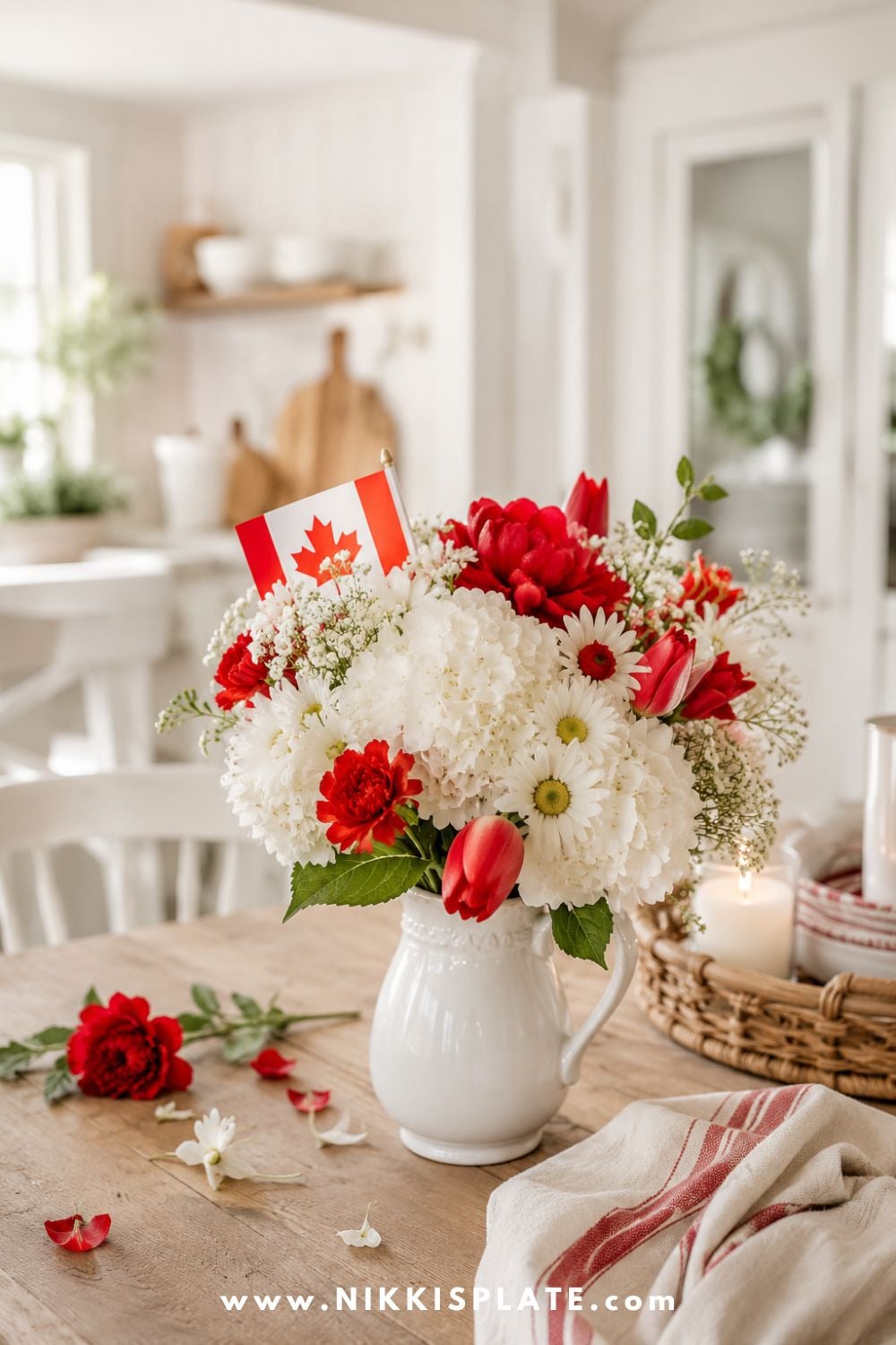 Canada Day flower arrangement with red and white blooms, mini Canadian flag, and a white pitcher styled on a rustic kitchen table.