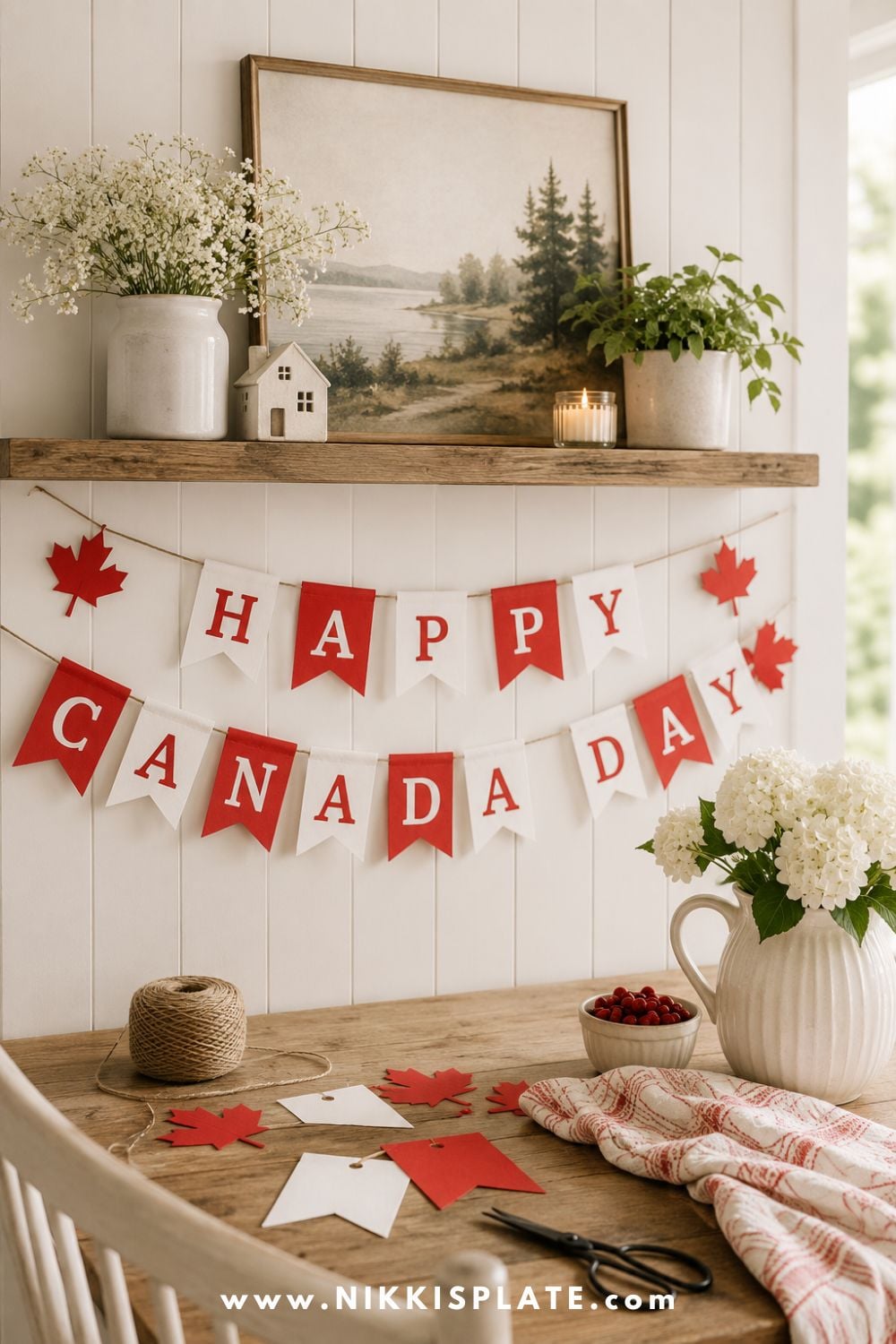 DIY Canada Day banner with red and white pennants, maple leaves, twine, flowers, and craft supplies styled on a rustic table beneath a mantel.