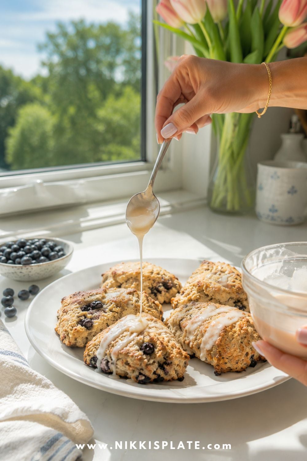 Female hand drizzling lemon glaze over freshly baked blueberry chia sourdough scones in a bright farmhouse kitchen.
