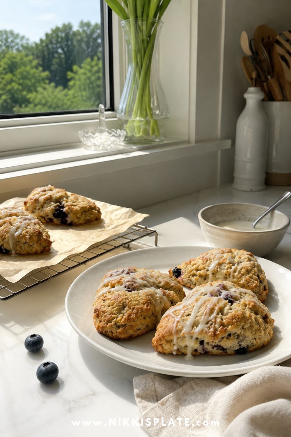Fresh blueberry chia sourdough scones with lemon glaze arranged on a white plate beside a cooling rack in a sunlit kitchen.