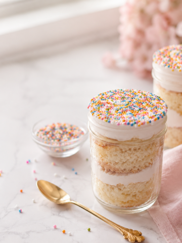 Mason jar dot cake recipe with visible layers of vanilla cake and white frosting, topped with rainbow dot sprinkles for a fun party dessert.
